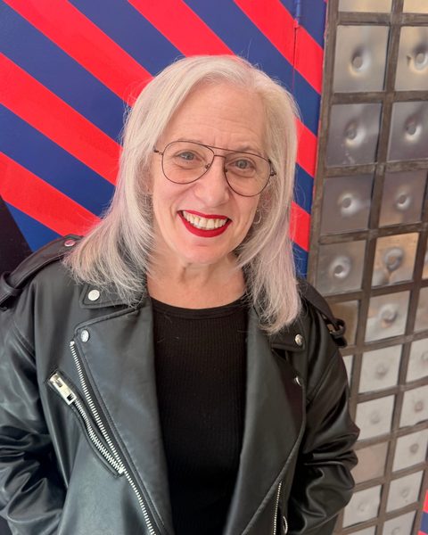 Suzanne Noble smiling in a black leather jacket in front of a red and blue striped mural
